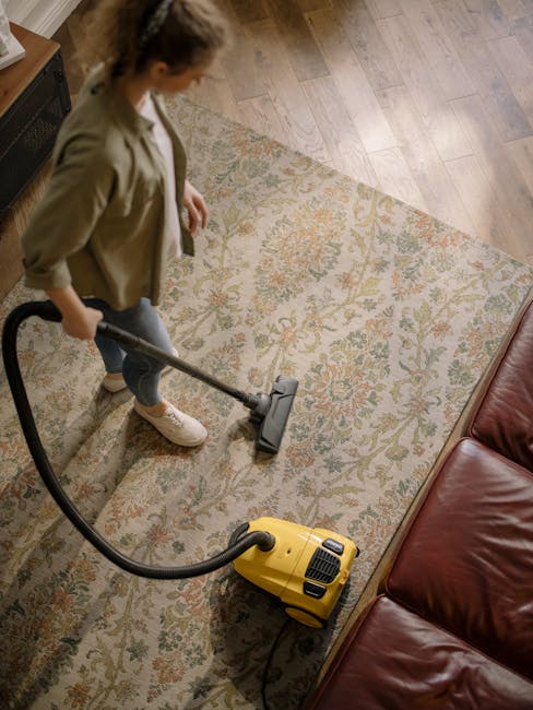 A woman engaged in surface cleaning with a yellow vacuum cleaner in a living room. She is wearing a beige jacket, dark pants, and white sneakers. The room features a patterned, beige and pastel floral rug on a light wooden floor, with a red leather sofa visible at the edge. The lighting is natural and bright, highlighting the clean, dust-free surface of the rug. This scene illustrates professional domestic cleaning services provided by Bayswater Carpet Cleaning, emphasizing deep cleaning and sanitization of residential carpets and floors.