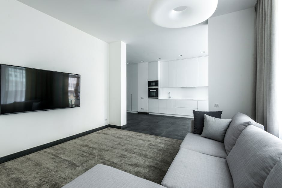 A modern, minimalistic living room with white walls and a light gray sofa adorned with two dark and light gray cushions. The room features a textured beige carpet covering the floor area in front of the sofa. On the left wall, there is a large flat-screen television mounted at eye level. The background reveals an open-plan kitchen with white cabinetry, integrated oven, and a stainless steel sink, set against dark flooring that contrasts with the lighter living space. Natural light filters in through a window with sheer white curtains on the right, illuminating the space evenly. The ceiling has a sleek, round, white light fixture. The room appears clean and well-maintained, highlighting the importance of surface cleaning and regular domestic cleaning handled by Bayswater Carpet Cleaning.