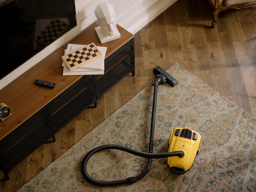A home living room featuring a wooden floor with a patterned area rug. A yellow vacuum cleaner with a black hose is placed on the rug, indicating surface cleaning activity. Against the wall, there is a black metal and wood media console topped with decorative items including a lamp, a bust sculpture, and a chessboard. A remote control rests on the console, and a white wall with molding is in the background, illuminated by soft natural light. Bayswater Carpet Cleaning specializes in deep cleaning and sanitisation for domestic and commercial spaces, as reflected in the well-maintained, clean appearance of the room.