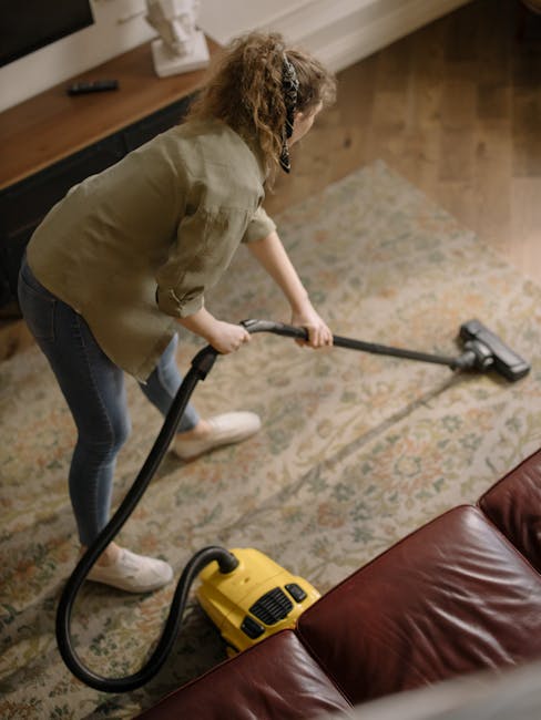 A woman with curly hair wearing a beige jacket, dark jeans, and white shoes is performing surface cleaning with a vacuum cleaner on a traditional patterned area rug within a living room. The vacuum cleaner, which is yellow with black components, is being used to deep clean the carpet, removing dust and debris. The room features a wooden sideboard with decorative items against the wall, and natural light illuminates the space, highlighting the cleanliness and well-maintained condition of the surfaces. The scene depicts a routine domestic cleaning process, emphasizing hygiene and maintenance in a residential setting, consistent with services offered by Bayswater Carpet Cleaning near Queensway station.