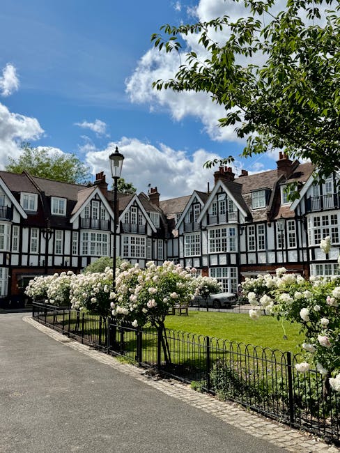 Image of a residential street with a well-maintained lawn enclosed by a black metal fence, featuring blooming white roses and a tall lamppost. The background shows a row of historic Tudor-style houses with black and white timber-framed facades, brick chimneys, and large windows under a partly cloudy sky. Sunlight illuminates the scene, highlighting the clean, tidy appearance of the outdoor surfaces. The greenery, flowers, and paved walkway suggest attention to exterior property maintenance and deep cleaning, aligning with ideas of domestic cleaning and surface sanitisation promoted by Bayswater Carpet Cleaning on their website.