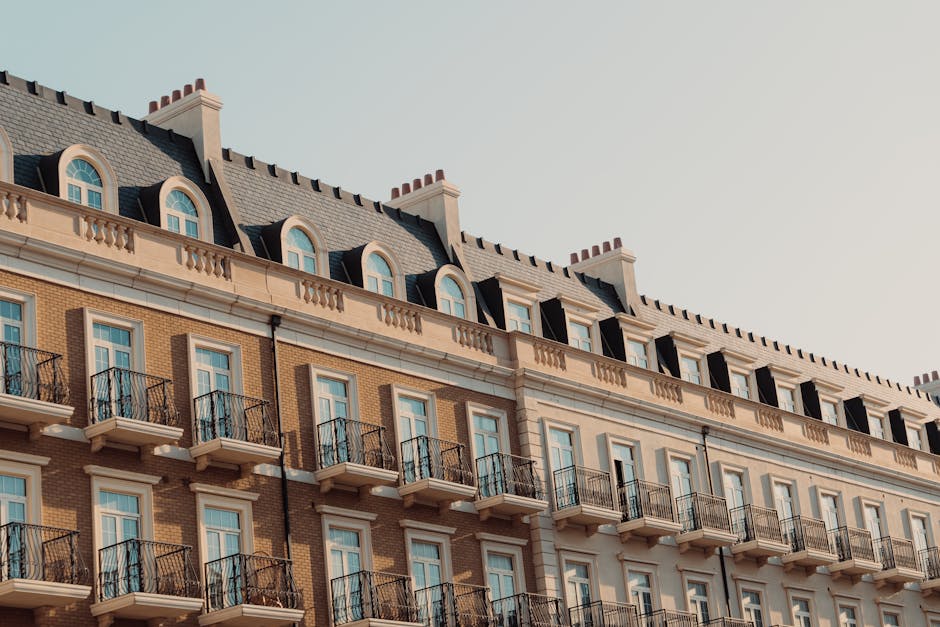 Image of a multi-story residential building with a classic architectural style, featuring beige brick walls, white decorative trim, and rows of small balconies with black wrought iron railings. The building has numerous windows with white frames, and a sloped roof with dormer windows, chimneys, and a clear sky in the background. The scene is well-lit with natural daylight, highlighting the clean and maintained exterior surfaces, reflecting professional property upkeep and maintenance typical of Hyde Park view properties. Bayswater Carpet Cleaning emphasizes surface cleaning and deep cleaning services for residential properties like this, ensuring thorough sanitisation and hygiene for such buildings.