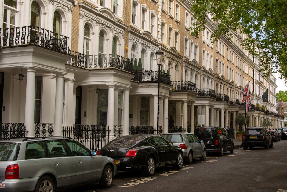 A row of elegant Victorian-style terraced houses in Bayswater, London, featuring white facades with decorative moldings and black wrought iron railings. The street is lined with parked cars, including a silver estate, a black coupe, and several SUVs. The pavement is clean and well-maintained, with lush green trees partially shading the street, and a British flag hanging from one of the buildings. The scene is brightly lit by natural daylight, highlighting the pristine condition of the exterior surfaces, reflective windows, and tidy surroundings, consistent with professional domestic cleaning and surface maintenance standards promoted by Bayswater Carpet Cleaning.